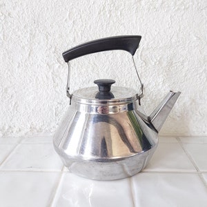 May include: A stainless steel tea kettle with a black handle and lid knob. The kettle has a spout for pouring and a hinged handle. The kettle is sitting on a white tiled surface.