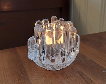 May include: A clear glass candle holder with a unique design featuring multiple vertical spikes. The candle holder is lit and casting a warm glow.