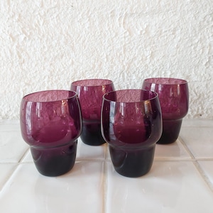 May include: Set of four purple glass tumblers. The glasses are clear with a deep purple hue and have a rounded shape.