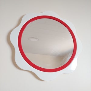 以下が含まれることがあります： 白い壁に掛けられた、白と赤の丸い鏡。鏡は波打つような縁取りがされている。