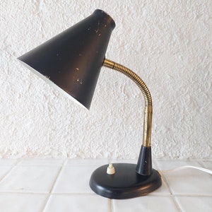 Puede incluir: Una lámpara de escritorio negra y dorada con un brazo flexible y una pantalla cónica negra. La lámpara tiene un interruptor blanco en la base.