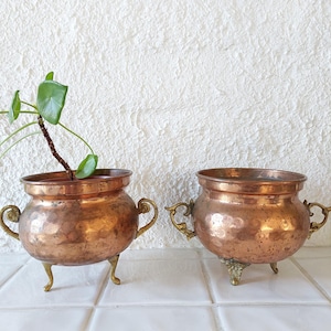 Puede incluir: Dos macetas de cobre martillado con asas y patas de latón. Las macetas están sobre una superficie de azulejos blancos. Una maceta tiene una pequeña planta verde dentro.