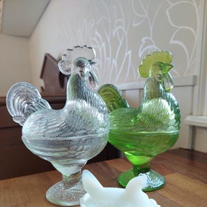 Puede incluir: Tres platos de dulces de vidrio con forma de gallo. Uno es transparente, otro verde y otro blanco.