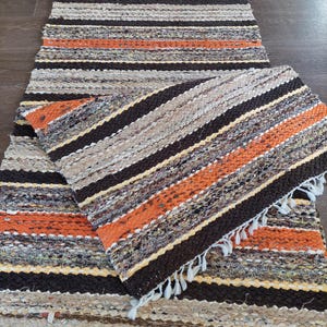 Könnte beinhalten: Ein gewebter Teppich mit braunen, orangen und beigefarbenen Streifen. Der Teppich hat einen Fransenrand und ist aus Naturfasern gefertigt.