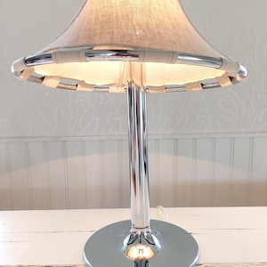 May include: A chrome table lamp with a beige fabric shade. The shade is trimmed with a light-colored woven material. The lamp has a round base and a tall, slender stem. The lamp is on a white surface.