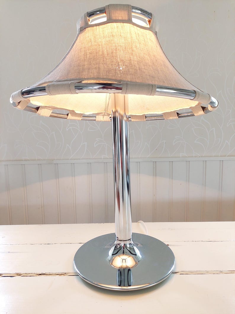 May include: A chrome table lamp with a beige fabric shade. The shade is trimmed with a light-colored woven material. The lamp has a round base and a tall, slender stem. The lamp is on a white surface.