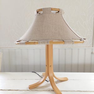 Może przedstawiać: Drewniana lampa stołowa z abażurem z naturalnego lnu. Lampa ma unikalną, tr&oacute;jnożną podstawę o zakrzywionym designie.