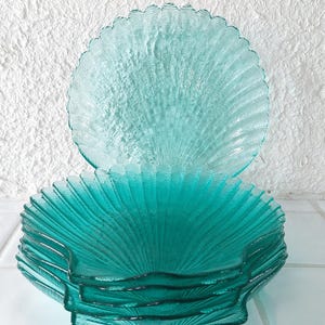 Op de afbeelding: Een stapel turquoise glazen borden met een geschulpte, schelpachtige vormgeving. De borden zijn op elkaar gestapeld, met één bord erbovenop. De achtergrond is een witte, getextureerde muur.