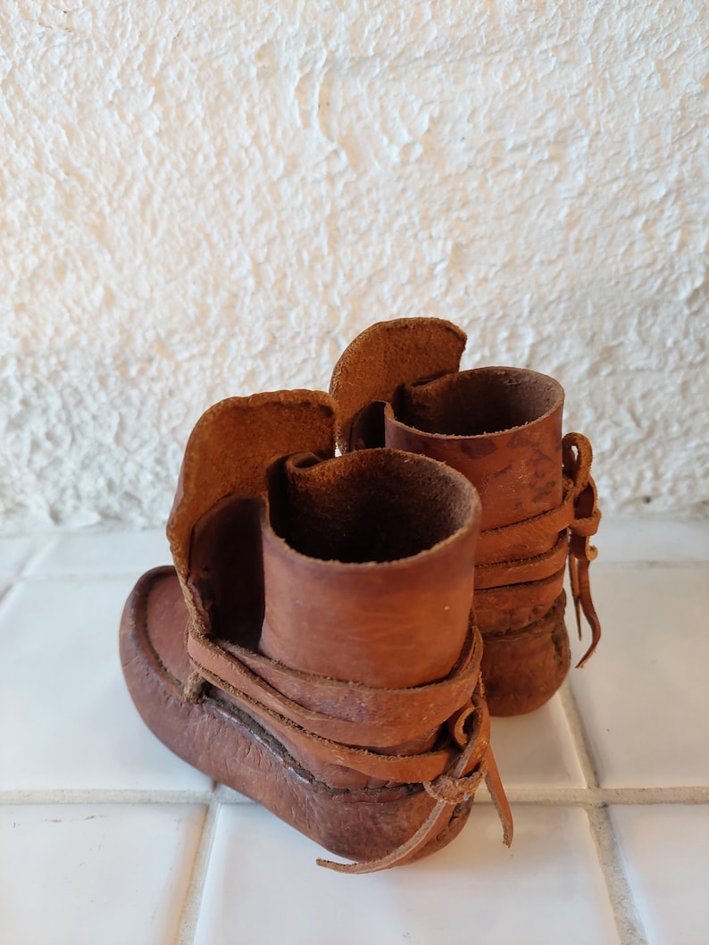 May include: A pair of brown leather moccasins with laces. The moccasins are tied at the ankle and have a fringe detail.