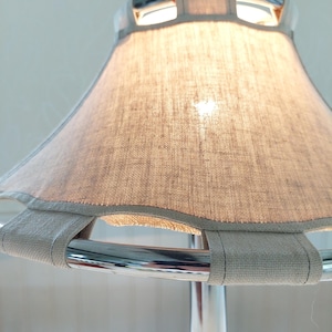 May include: A close-up of a modern lamp with a light gray linen shade. The shade is supported by a chrome frame and has a light gray trim. The lamp is turned on, and the light shines through the shade.