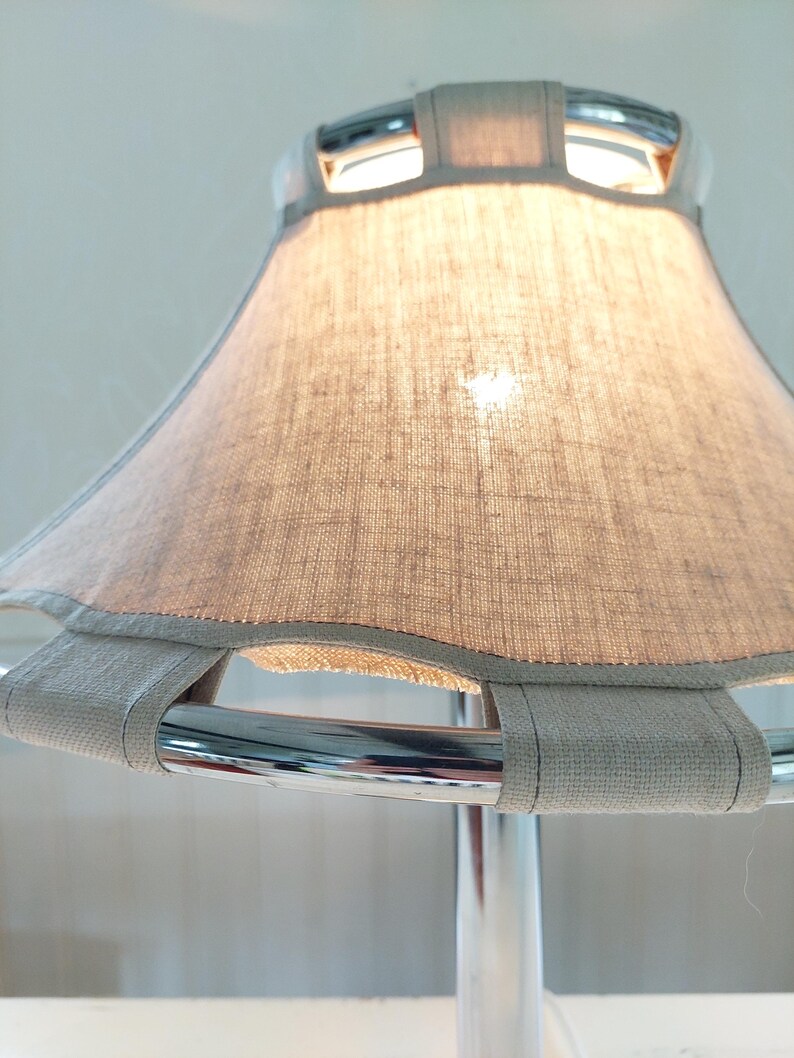 May include: A close-up of a modern lamp with a light gray linen shade. The shade is supported by a chrome frame and has a light gray trim. The lamp is turned on, and the light shines through the shade.