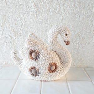 Może przedstawiać: Dekoracyjna rzeźba łabędzia wykonana z małych, białych muszli. Łabędź ma trzy akcenty kwiatowe z muszli na ciele oraz szczegółową głowę i szyję. Rzeźba jest ustawiona na białym, teksturowanym tle.