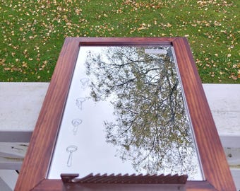 May include: A brown wooden framed mirror with a silver reflective surface. The mirror reflects a tree with green leaves and a cloudy sky. There are several pencil drawings on the mirror surface.
