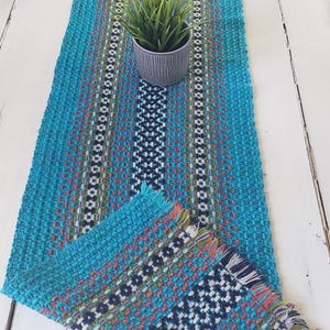 May include: A woven turquoise table runner with a geometric pattern in white, black, and orange. The runner has fringed edges and a central stripe. A small potted plant sits on the runner.