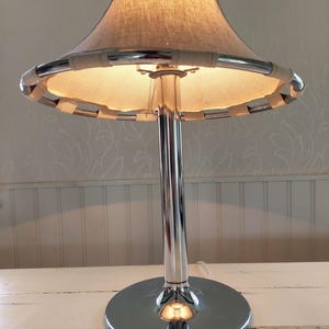 May include: A chrome table lamp with a beige fabric shade. The shade is supported by a chrome frame and has a decorative trim. The lamp has a round base and a slender, curved stem. The lamp is turned on, illuminating the shade.