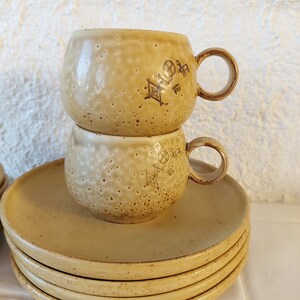 May include: A set of four yellow ceramic saucers with a stack of two yellow ceramic mugs with brown markings on top. The mugs have a rounded shape and a handle.