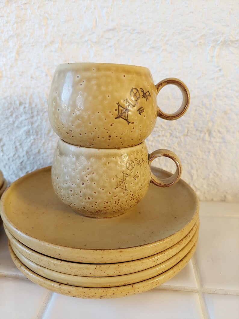 May include: A set of four yellow ceramic saucers with a stack of two yellow ceramic mugs with brown markings on top. The mugs have a rounded shape and a handle.