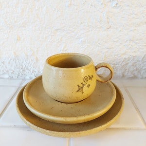 May include: A set of three ceramic dishes in a light yellow color with a speckled texture. The cup has a handle and a design on the side. The dishes are stacked on top of each other.