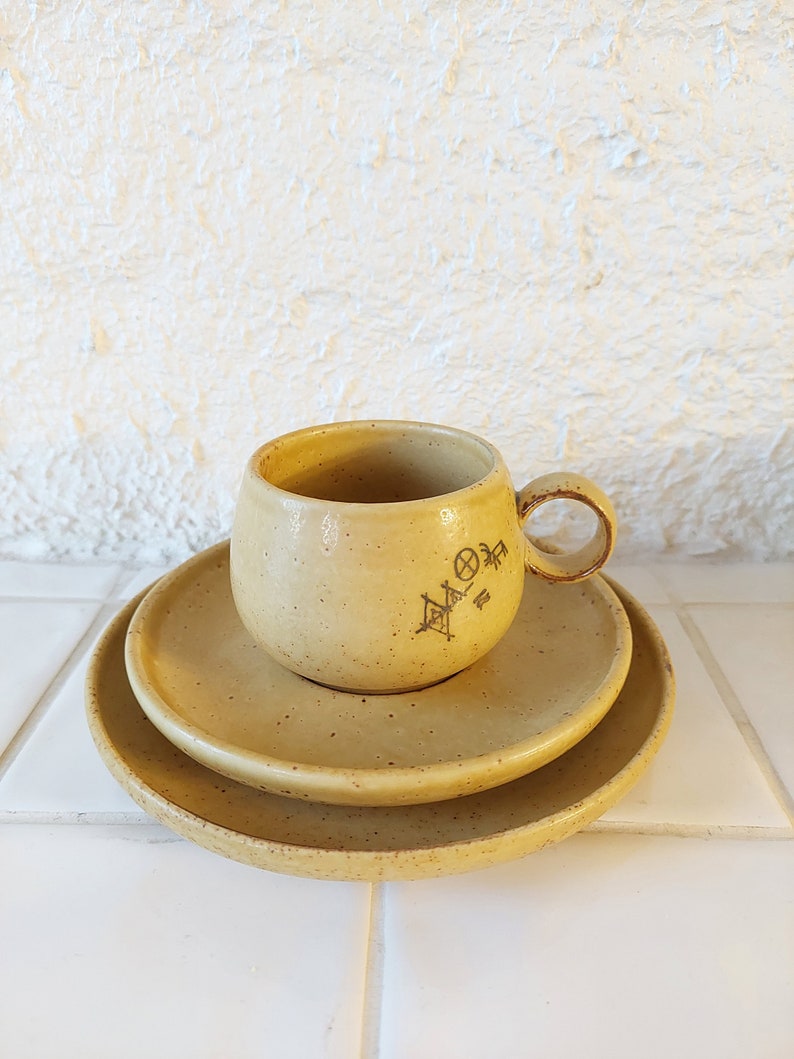 May include: A set of three ceramic dishes in a light yellow color with a speckled texture. The cup has a handle and a design on the side. The dishes are stacked on top of each other.