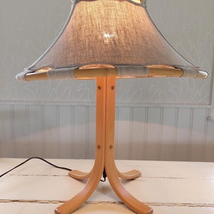 Może przedstawiać: Drewniana lampa stołowa z abażurem z jasnoszarego lnu. Abażur ma drewniany stelaż z wycięciami. Lampa ma tr&oacute;jnożną podstawę.