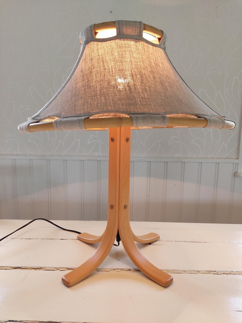 Może przedstawiać: Drewniana lampa stołowa z abażurem z jasnoszarego lnu. Abażur ma drewniany stelaż z wycięciami. Lampa ma tr&oacute;jnożną podstawę.