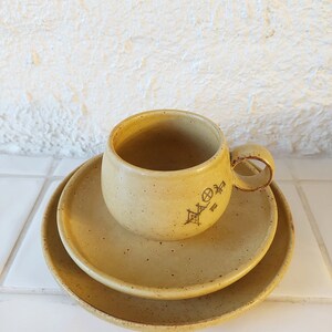 May include: A set of three ceramic dishes in a light yellow color with a speckled finish. The dishes are stacked with a cup on top. The cup has a handle and a symbol on the side.