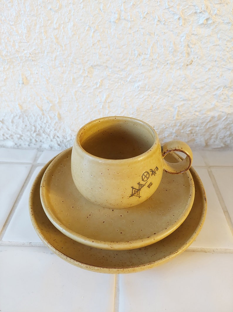 May include: A set of three ceramic dishes in a light yellow color with a speckled finish. The dishes are stacked with a cup on top. The cup has a handle and a symbol on the side.