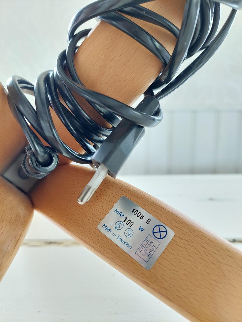 Może przedstawiać: Zbliżenie na drewnianą podstawę lampy z czarnym kablem elektrycznym i etykietą z napisem "Max 100 W 4008 B Wyprodukowano w Szwecji".