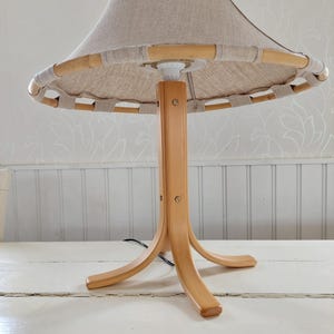 Może przedstawiać: Drewniana lampa stołowa z naturalnym abażurem lnianym i bambusową obw&oacute;dką. Lampa ma tr&oacute;jnożną podstawę.