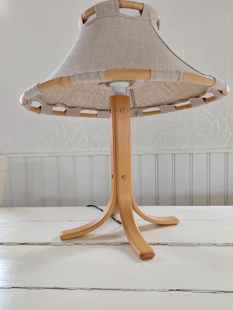 Może przedstawiać: Drewniana lampa stołowa z naturalnym abażurem lnianym i bambusową obw&oacute;dką. Lampa ma tr&oacute;jnożną podstawę.