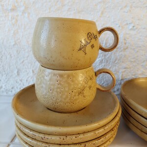 May include: A set of four beige ceramic saucers with a speckled pattern and a matching set of two beige ceramic mugs with a speckled pattern and a geometric design.
