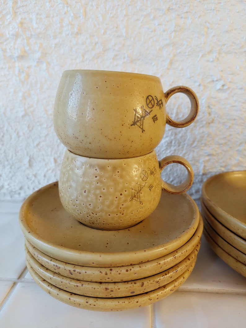 May include: A set of four beige ceramic saucers with a speckled pattern and a matching set of two beige ceramic mugs with a speckled pattern and a geometric design.