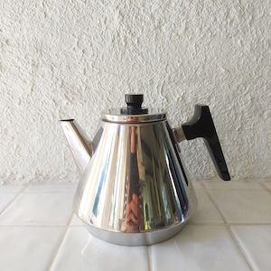 Könnte beinhalten: Eine silberne, kegelförmige Teekanne mit schwarzem Griff. Die Teekanne hat eine glänzende, reflektierende Oberfläche.