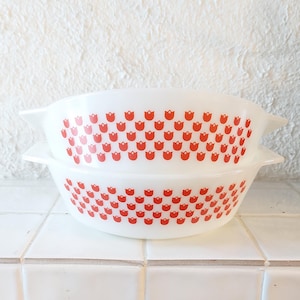 Op de afbeelding: Twee witte Pyrex-kommen met rode tulp patronen. De kommen zijn gestapeld, waarbij hun afgeronde vorm en zijhandvatten worden getoond. Het rode tulp ontwerp is gelijkmatig verdeeld over de buitenkant van de kommen.