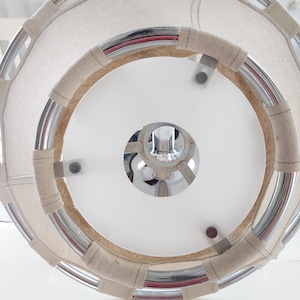 May include: A circular, modern light fixture with a white interior and a chrome frame. The frame is wrapped with a beige fabric. The fixture is suspended from the ceiling, showcasing a clean, contemporary design.