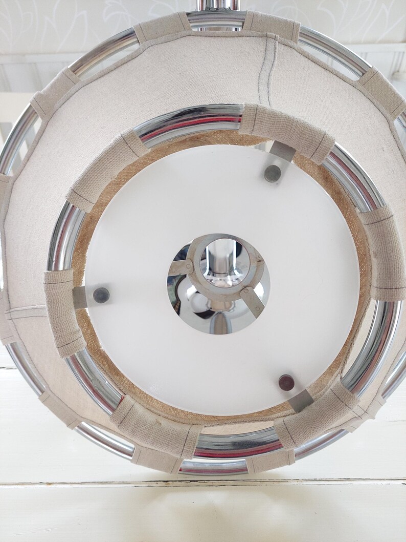May include: A circular, modern light fixture with a white interior and a chrome frame. The frame is wrapped with a beige fabric. The fixture is suspended from the ceiling, showcasing a clean, contemporary design.