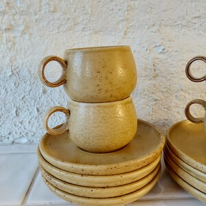 May include: A stack of six beige ceramic saucers with a speckled texture. Two matching beige ceramic cups are stacked on top of the saucers.