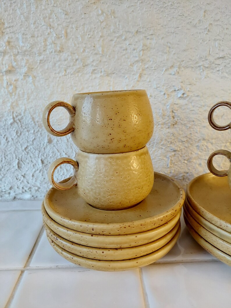 May include: A stack of six beige ceramic saucers with a speckled texture. Two matching beige ceramic cups are stacked on top of the saucers.