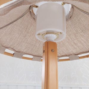 Może przedstawiać: Drewniana lampa stojąca z białym abażurem z tkaniny. Lampa ma prosty design i jest wykonana z naturalnych materiał&oacute;w.