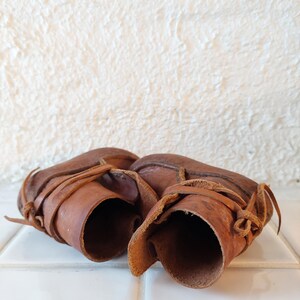May include: A pair of brown leather baby booties with laces. The booties are tied together and are sitting on a white tiled surface.