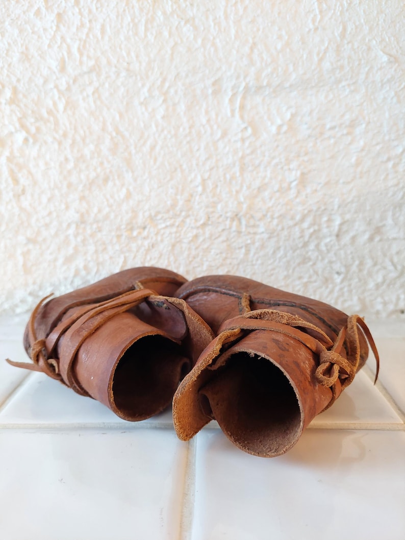 May include: A pair of brown leather baby booties with laces. The booties are tied together and are sitting on a white tiled surface.