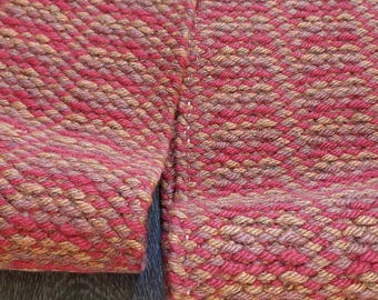 May include: Close-up of a section of woven fabric in shades of red, brown, and tan. The fabric has a textured, diagonal pattern. The edges are visible, showing the seam with white stitching. The background is a wooden surface.