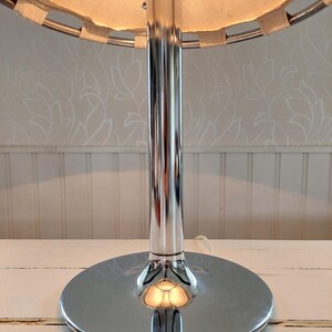 May include: A chrome table lamp with a circular base and a cylindrical stem. The lamp has a beige fabric shade with a woven trim. The lamp is on a white surface.