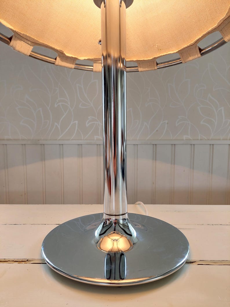 May include: A chrome table lamp with a circular base and a cylindrical stem. The lamp has a beige fabric shade with a woven trim. The lamp is on a white surface.
