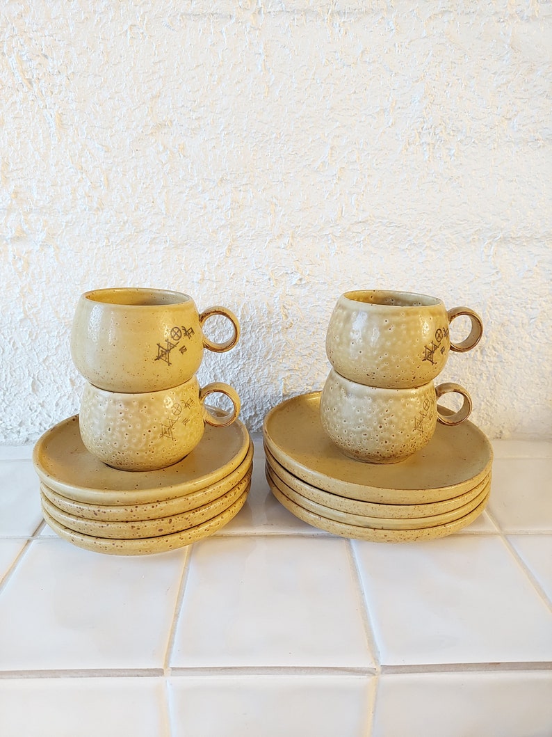 May include: Two sets of stacked ceramic coffee cups with matching saucers. The cups and saucers are a speckled beige color with a brown design.