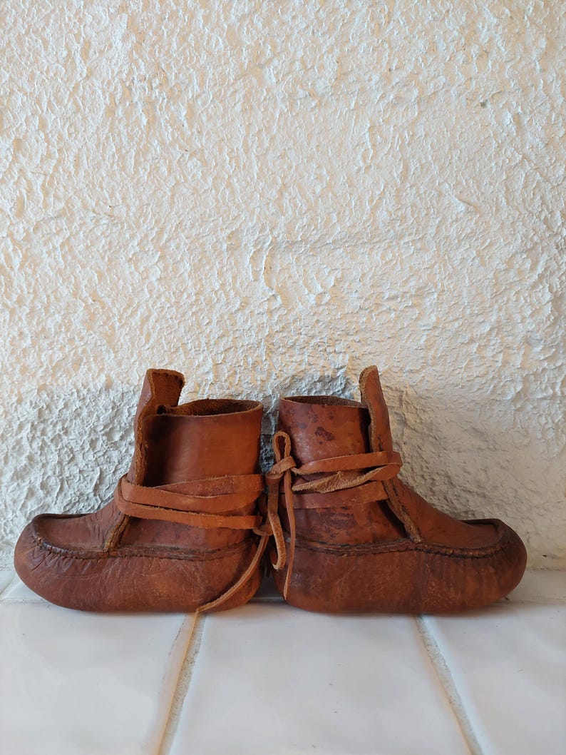 May include: A pair of brown leather moccasins with laces. The moccasins are tied together and have a worn look.