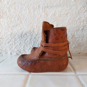 May include: A pair of brown leather baby booties with a strap and buckle closure. The booties are made of a soft, supple leather and have a slightly pointed toe.