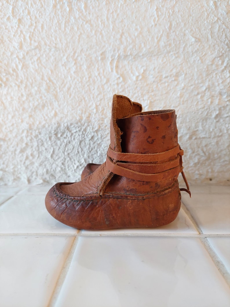 May include: A pair of brown leather baby booties with a strap and buckle closure. The booties are made of a soft, supple leather and have a slightly pointed toe.