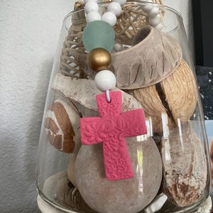 May include: A clear glass vase filled with decorative items, including seashells and a pink cross pendant. The pendant is strung with white, gold, and green beads. The vase sits on a white decorative base.