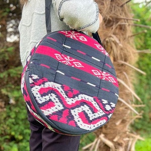 Puede incluir: Un bolso de hombro redondo y acolchado con una correa negra. El bolso presenta un patrón geométrico en rojo, blanco y negro. El bolso se lleva sobre el hombro.